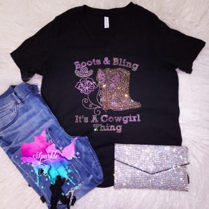 May include: Black V-neck t-shirt with rhinestone design reading "Boots & Bling" and "It's A Cowgirl Thing". Includes blue jeans with a colorful paint splatter design and a sparkly silver clutch.