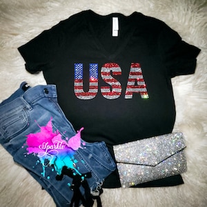 May include: A black V-neck t-shirt featuring the text USA in a red, white, and blue rhinestone flag pattern. The shirt is paired with blue denim jeans and a silver rhinestone-covered envelope clutch bag, creating a coordinated ensemble.