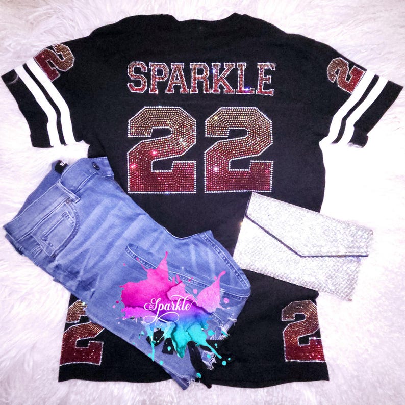 Puede incluir: Jersey negro con rayas blancas en las mangas. El jersey tiene la palabra "SPARKLE" y el n&uacute;mero "22" en pedrer&iacute;a. La pedrer&iacute;a es de color rojo y dorado.