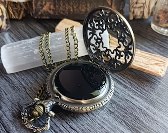 May include: An antique-style bronze pocket watch with a smooth black circular face and an ornate, filigree-patterned hinged cover. The watch is attached to a matching bronze link chain, featuring intricate decorative metalwork on the winding crown.