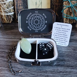 May include: A silver-toned metal case containing a light green oval stone pendant on a chain, three black dice with white pips, and a white card titled Dice Roll Divination. The lid interior features a circular zodiac chart with text.