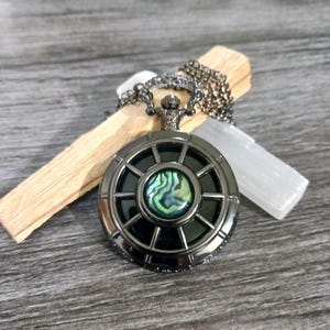 May include: A dark gray pendant necklace with a round, wheel-like design. The center features an iridescent, abalone-like stone. The necklace is displayed with a piece of wood and a white crystal, suggesting a spiritual or bohemian theme.