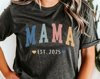 May include: A dark gray t-shirt with the word "MAMA" in colorful letters. The letters are in a rainbow color scheme. Below the word "MAMA" is the text "EST. 2025" with a small heart on either side.