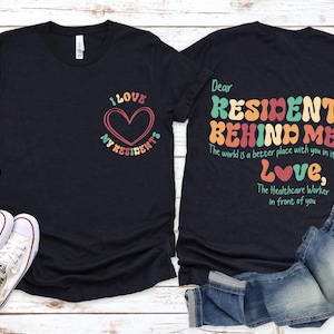 May include: Two black t-shirts with a colorful graphic design. The front shirt says "I love my residents" with a heart. The back shirt says "Dear Resident Behind Me, The world is a better place with you in it! Love, The Healthcare Worker in front of you".