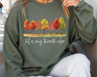 May include: Olive green sweatshirt featuring a watercolor design of colorful autumn trees. The trees display shades of red, orange, and yellow. The text "fall is my favorite color" is written in a cursive font below the trees.