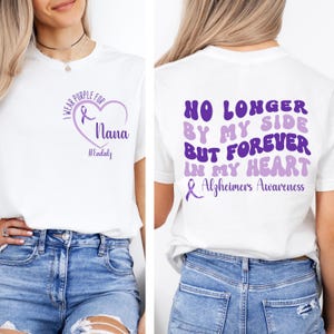 May include: White t-shirt with purple text and graphics. The front features a heart with a ribbon, the words "I Wear Purple For Nana", and "HEndalz". The back says "No Longer By My Side But Forever In My Heart Alzheimer's Awareness".