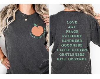 May include: A dark gray t-shirt with a peach graphic and the text "Galations 5:22-23" on the front. The back of the shirt has the text "Love, Joy, Peace, Patience, Kindness, Goodness, Faithfulness, Gentleness, Self Control" printed in green.