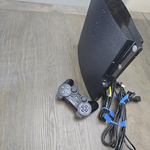 May include: Black PlayStation 3 console with a matching controller and power cables. The console is angled, showing its sleek design. The controller is gray with the Sony logo.