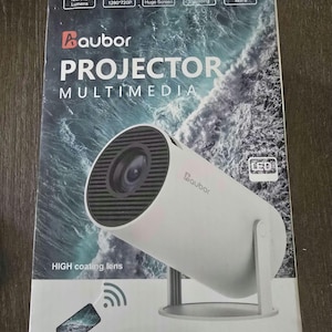 Op de afbeelding: Een witte Baubor multimedia projector met een zwarte lens, ondersteund door een standaard. De doos toont specificaties waaronder 120 ANSI lumen, 720P resolutie en tot 330 cm schermgrootte. De tekst "HIGH coating lens" is zichtbaar.