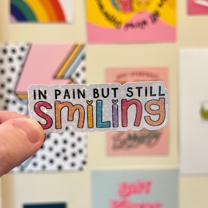 May include: A colourful sticker with the text "In pain but still smiling" in rainbow letters with glitter accents.