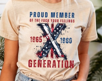 May include: A beige t-shirt with a graphic design featuring a red, white and blue American flag with a large X in the centre. The text "Proud Member of the Fuck Your Feelings 1965 - 1980 Generation" is printed on the shirt.