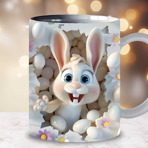 May include: White ceramic mug with a colorful image of a cartoon bunny rabbit peeking out of a nest of white eggs. The bunny has blue eyes and a big smile. The mug has a black rim and a white handle.