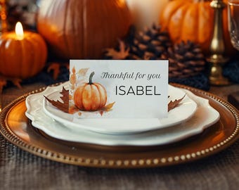 May include: A Thanksgiving table setting with a place card that reads "Thankful for you ISABEL" next to a watercolor pumpkin illustration. The table includes white plates, a gold charger, and a lit candle.