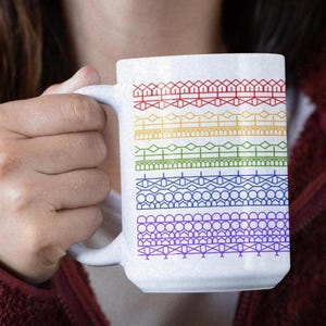 May include: White ceramic mug featuring a rainbow of geometric patterns. The mug's design includes rows of red, orange, yellow, green, blue, and purple shapes. The mug has a large handle and is held by a person.