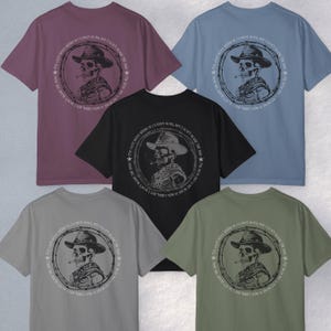 May include: A collection of five t-shirts in various colors: mauve, black, light blue, grey, and olive green. Each shirt features a circular graphic of a skeleton wearing a cowboy hat and smoking a cigarette, with text around the edge.