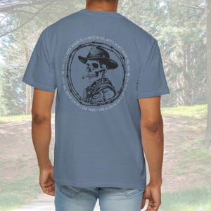 May include: A blue t-shirt featuring a circular graphic of a skeleton wearing a hat and smoking a cigarette. The graphic is surrounded by barbed wire and text. The text reads "I've got seven weeks in county bots, but I don't blame the man."