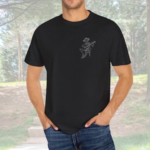 May include: Black t-shirt featuring a small, gray graphic of a skeleton playing a guitar and wearing a cowboy hat. The design is on the left chest area. The shirt is a solid black color and has short sleeves.