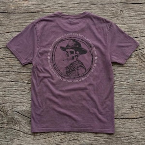 May include: A purple t-shirt with a graphic of a skeleton wearing a cowboy hat and smoking a cigarette. The design is encircled by text that reads "I've got some poser chips, but I won't blame the mix. I'm not born in county, but I don't blame the mix."