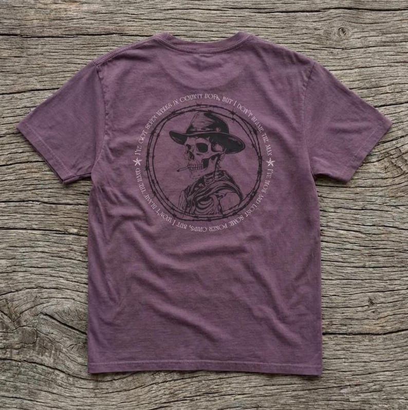 May include: A purple t-shirt with a graphic of a skeleton wearing a cowboy hat and smoking a cigarette. The design is encircled by text that reads "I've got some poser chips, but I won't blame the mix. I'm not born in county, but I don't blame the mix."