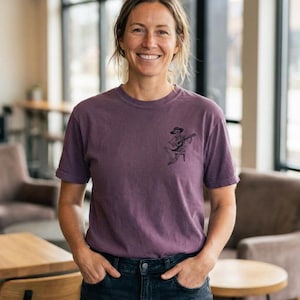 May include: A purple t-shirt with a crew neck and short sleeves. The shirt features a small black graphic of a skeleton playing a guitar. The person is wearing dark blue jeans and is smiling.