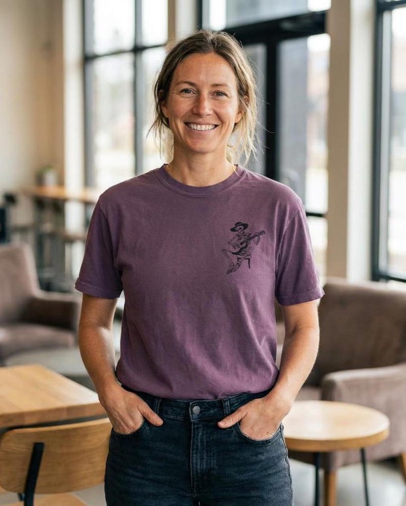 May include: A purple t-shirt with a crew neck and short sleeves. The shirt features a small black graphic of a skeleton playing a guitar. The person is wearing dark blue jeans and is smiling.