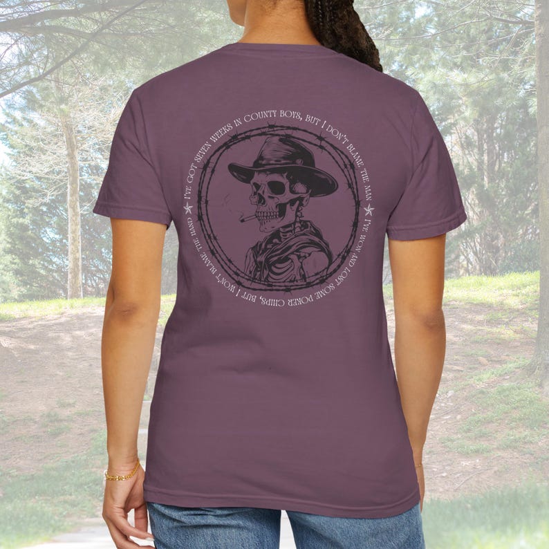 May include: Mauve t-shirt featuring a circular graphic of a skeleton wearing a cowboy hat and smoking a cigarette. The graphic is surrounded by text that reads "I've got seven weeks in county boys, but I don't blame the man."