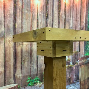May include: A handcrafted wooden pedestal table with a square top and a sturdy, rectangular base. The natural wood grain is visible, and the table is constructed with visible screws and dowels. The background features a wooden fence and string lights.