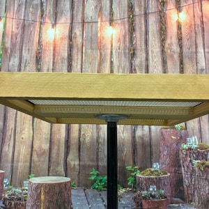 May include: A square wooden table with a metal mesh top and a black metal pipe leg. The table is supported by a wooden base and is set against a wooden fence backdrop. Decorative lights are strung above.