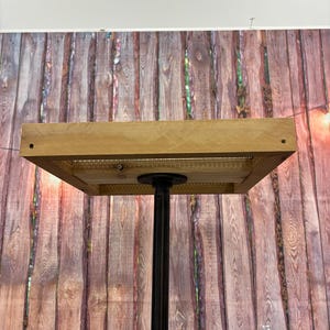 May include: A square, light-colored wooden table top with a metal mesh insert is supported by a black metal pipe and a wooden base. The table is designed for display purposes.