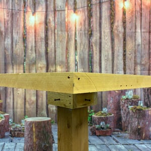 May include: A partially constructed wooden table with a light brown finish. The table features a square top and a single, sturdy leg. The background includes a wooden fence, string lights, and decorative tree stumps.