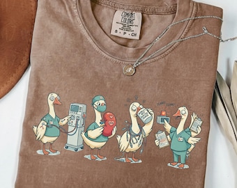 May include: Brown Comfort Colors t-shirt featuring a cartoon of geese dressed as medical professionals. The geese are depicted with medical equipment and holding items like a kidney and a coffee mug that says "World's Best Dialysis Nurse".