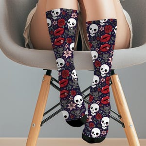 Peut inclure: Une paire de chaussettes hautes, avec un fond violet foncé. Les chaussettes présentent un motif répété de crânes blancs, de lèvres rouges, de chauves-souris grises, de fleurs roses et de roses rouges. Les chaussettes sont portées par une personne assise sur une chaise.