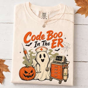 May include: A cream-colored t-shirt featuring a graphic of a ghost wearing a nurse cap and stethoscope, a jack-o'-lantern, a syringe, and a medical cart. The text reads Code Boo In The ER, accompanied by illustrations of bats and a spider web.