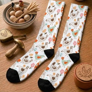 May include: A pair of white crew socks featuring a repeating pattern of brown, orange, and teal chickens interspersed with small flowers. The socks have solid black heels and toes, and are displayed alongside a bowl of eggs and a wooden stamp.