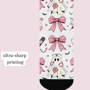 May include: A white sock featuring a repeating pattern of ghosts dressed as nurses, pink bows, medical supplies, and small bats. The ghosts wear nurse caps with red crosses and carry medical bags. The sock has a solid black toe section.
