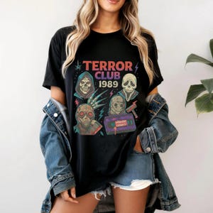 以下が含まれることがあります： 「TERROR CLUB 1989」の文字と、頭蓋骨やVHSテープなど、ホラーをテーマにしたイラストが描かれた黒いTシャツ。デザインは赤、紫、青の色合いです。