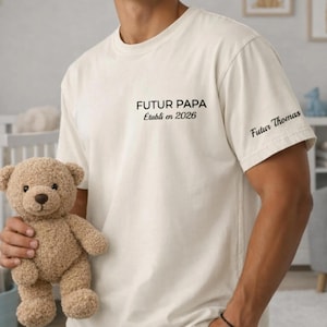 Puede incluir: Camiseta color crema con el texto "FUTUR PAPA Établi en 2026" impreso en el pecho y "Futur Thomas" en la manga. Un osito de peluche marrón se sostiene en la imagen. La camiseta es de estilo casual.