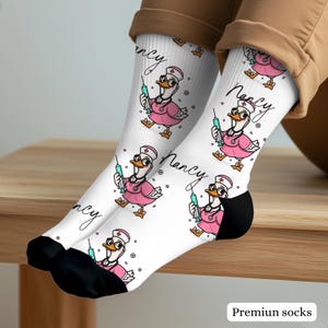 May include: White crew socks featuring a repeating pattern of a cartoon duck dressed as a nurse in a pink uniform with a stethoscope and a syringe. The name Nancy is written in black cursive script next to each duck. The socks have black toe sections.