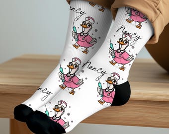 May include: White crew socks featuring a repeating pattern of a cartoon duck dressed as a nurse in a pink uniform with a stethoscope and a syringe. The name Nancy is written in black cursive script next to each duck. The socks have black toe sections.