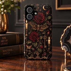 May include: A black smartphone case featuring a detailed print of deep red roses, a moth with skull markings, two lit candles in gold holders, a gold skeleton key, and a crescent moon against a dark background with ornate gold filigree accents.