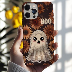 May include: A phone case featuring a cartoon ghost with a lace veil, surrounded by brown sunflowers on a dark brown background. The word "BOO" is displayed in white letters. The case is held in a hand.