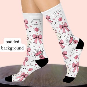 May include: White crew socks featuring a repeating pattern of pink bows, medical caps with red crosses, pink face masks, syringes with spider webs, small pumpkins, pills, and tiny black bats. The socks have solid black heels and toe sections.