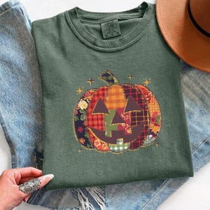 May include: A dark olive green crew neck t-shirt featuring a central graphic of a pumpkin composed of various fabric-like patches in plaid, floral, and checkered patterns. The pumpkin has a dark brown jack-o'-lantern face and small gold stars.