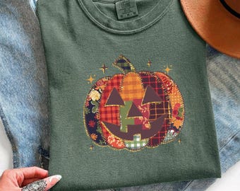 Op de afbeelding: Een donker olijfgroen t-shirt met ronde hals en een centrale afbeelding van een pompoen die is samengesteld uit diverse stofachtige lapjes met ruitjes-, bloemen- en geblokte patronen. De pompoen heeft een donkerbruin gezicht en gouden sterren.