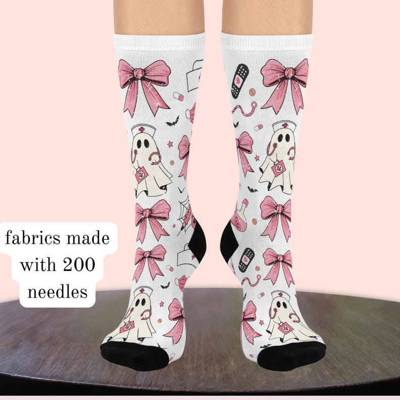 May include: White crew socks featuring a repeating pattern of pink bows, ghost figures wearing nurse caps and stethoscopes, bandages, and small bats. The socks include text stating fabrics made with 200 needles and have black toe sections.