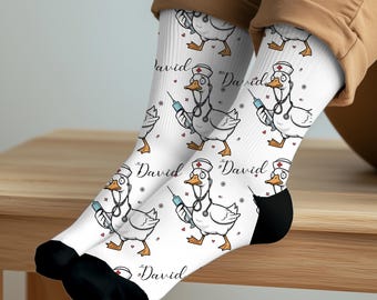 May include: White crew socks featuring a repeating cartoon pattern of a white duck dressed as a nurse, wearing a cap with a red cross and a stethoscope, holding a blue syringe. The name David is printed in black script between the duck illustrations.