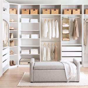Könnte beinhalten: Ein großes weißes modulares Schranksystem mit offenen Regalen, Kleiderstangen und Schubladen. Die Regale enthalten geflochtene Körbe und gefaltete weiße Wäsche. Eine hellgraue gepolsterte Aufbewahrungsbank mit einer weißen Decke steht davor.