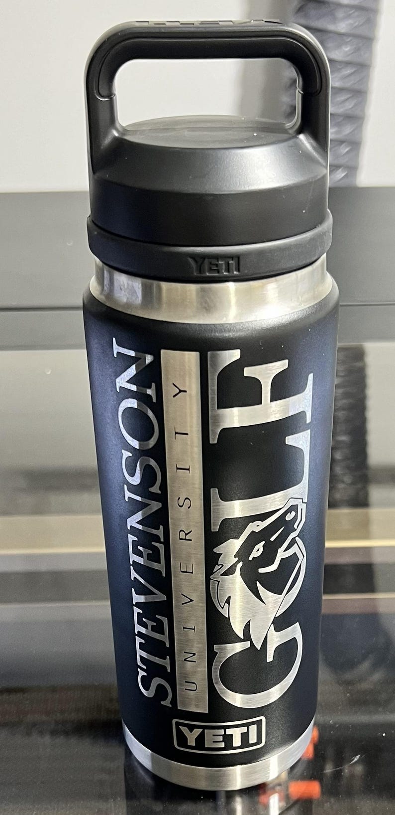May include: Black insulated water bottle with a handle, featuring the text "STEVENSON UNIVERSITY GOLF" in silver lettering. The bottle has a silver band and the brand name "YETI" is visible.