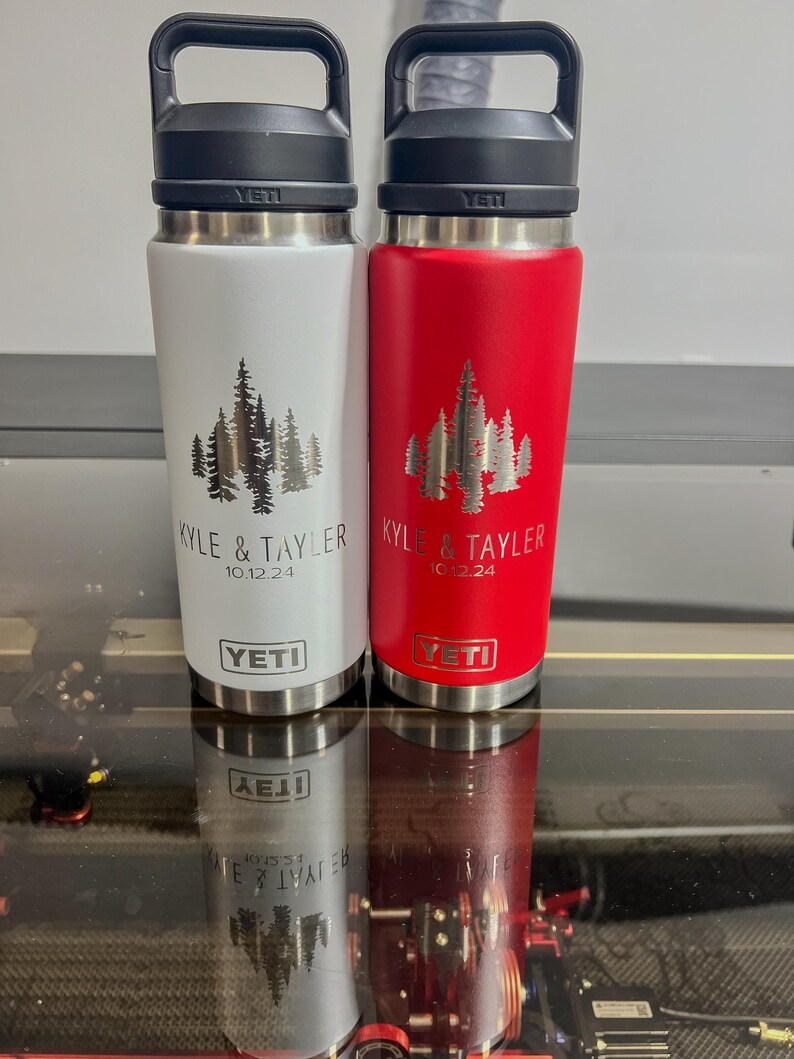 May include: Two stainless steel Yeti water bottles, one white and one red, with black handles. Each bottle features a forest design and the names "KYLE & TAYLER" with the date "10.12.24". The Yeti logo is visible.