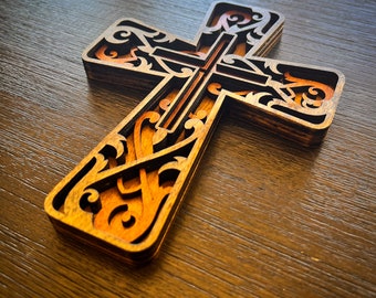 May include: A layered wooden cross with intricate, swirling cut-out designs. The cross features a warm, natural wood tone with a contrasting orange interior, creating a three-dimensional effect. The design is detailed and ornate, suitable for religious decor.